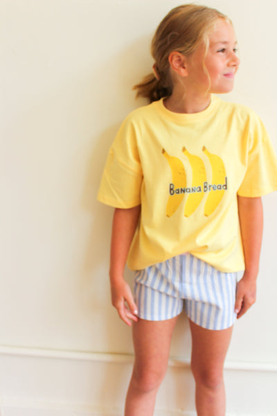 a yellow 'Banana Bread' t-shirt and striped shorts against a white wall.