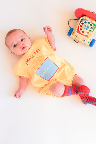 Baby wearing a yellow onesie with small fry graphic lying on a white surface next to a toy phone.