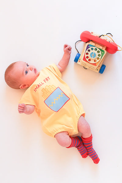 Baby lying on a white surface wearing a yellow romper with small fry graphic 