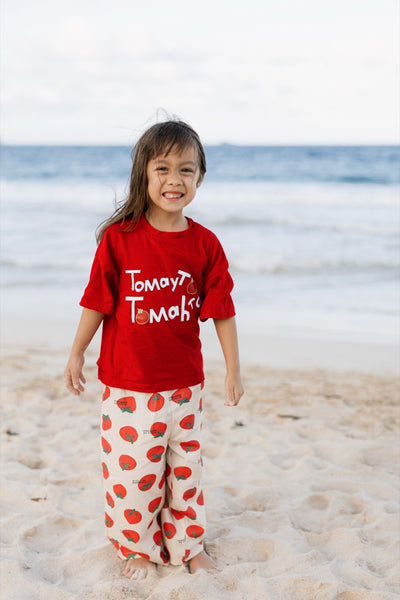Child wearing a red shirt and white pants with red tomato patterns on a beach