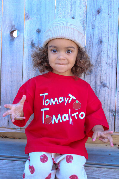 A toddler girl wearing a bright red short-sleeve T-shirt with white text reading 'Tomayto tomahto' graphic and white pants with red tomato prints