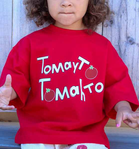 A child wearing a bright red short-sleeve T-shirt with white text reading 'Tomayto tomahto' graphic and white pants with red tomato prints
