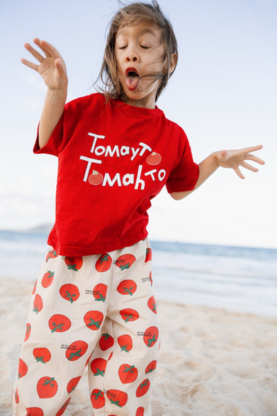 Child wearing a red 'Tomato Tomato' shirt and white pants with tomato pattern on a beach.