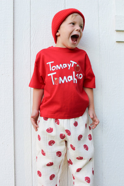 A young child wearing a red beanie, a red t-shirt with 'Tomat Tomatko' in white text, and cream-colored pants with red tomato prints, standing against a white paneled wall.