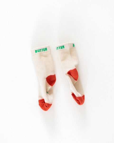 A pair of beige socks with the word 'BUTTER' printed in green on the top, designed for kids.