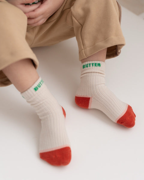 A pair of beige socks with the word 'BUTTER' printed in green on the top, designed for kids.