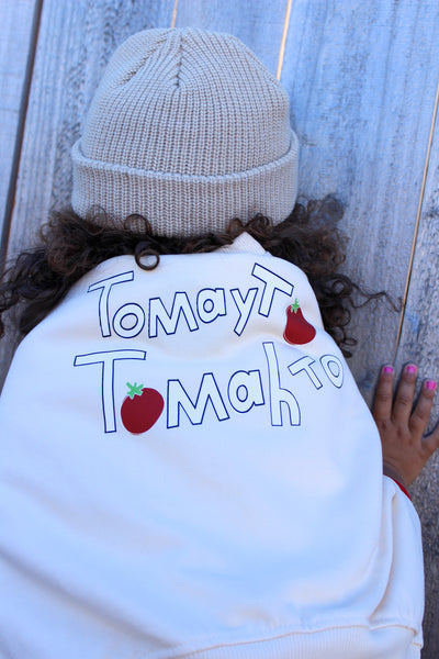 A child seen from behind wearing a white sweatshirt with a humorous tomato-themed graphic spelling 'Tomayto Tomahto' and red tomato illustrations, paired with a beige knit beanie and curly brown hair, leaning against a weathered gray wooden wall.