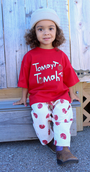 A young child wearing a red graphic t-shirt with 'Tomato' written on it, white pants with red tomato prints, and a cream knit beanie, sitting on a wooden bench against a weathered wood fence.