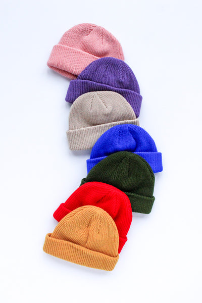 Six knit beanie hats in different colors are arranged diagonally on a white background. The colors are pink, purple, beige, blue, dark green, and mustard yellow.