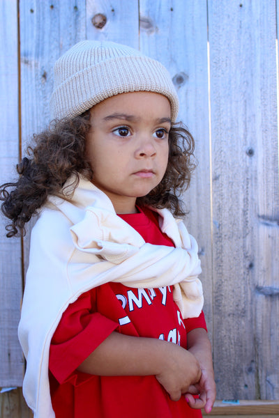 A child wearing a bright red short-sleeve T-shirt with white text reading 'Tomayto tomahto' graphic and white pants with red tomato prints  and a white beanie 