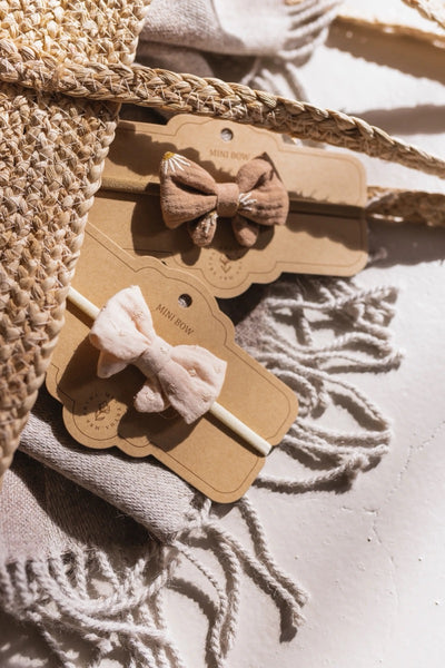 A headband with a beige cotton bow placed on a neutral-toned surface, likely cardboard packaging, with a textured background.