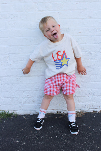 Child wearing a white t-shirt with a colorful graphic and red checkered shorts standing against a white brick wall.