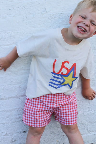 Child wearing a white t-shirt with 'USA' design and red checkered shorts against a white brick wall.
