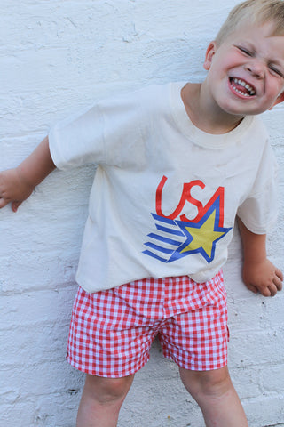 Child wearing a white t-shirt with 'USA' design and red checkered shorts against a white brick wall.