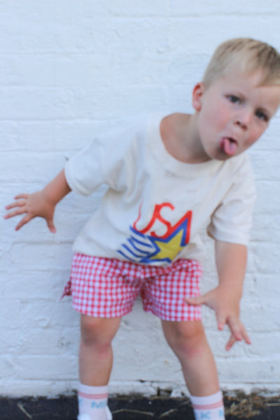 Child wearing a white sweatshirt with 'USA' print and red checkered shorts against a white brick wall.