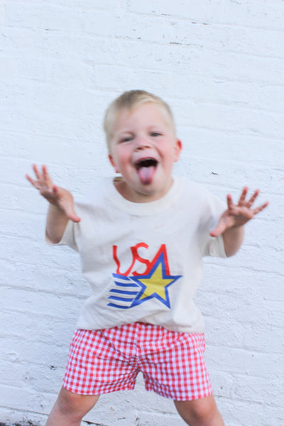 Child wearing a shirt with 'USA' and a star design, standing against a white brick wall.