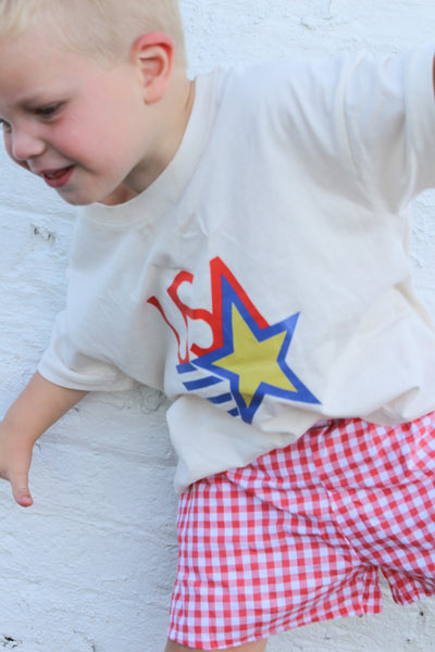 Child wearing a white shirt with a colorful USA graphic and red checkered shorts against a white brick wall.