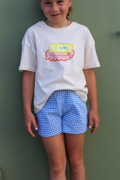 cream butter dish  t-shirt with a colorful design and blue checkered gingham shorts with red bow on side. 