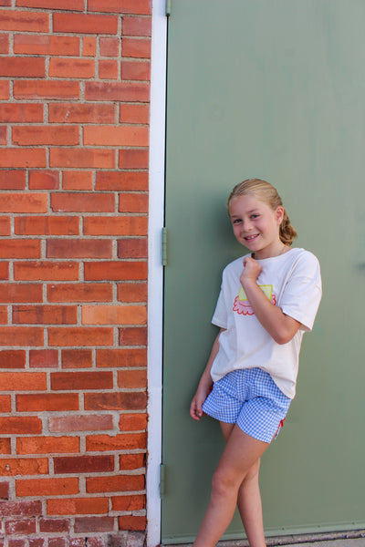 cream butter dish  t-shirt with a colorful design and blue checkered gingham shorts with red bow on side. 