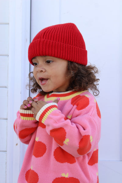 A young child wearing a bright red ribbed knit beanie and a pink sweater with large red apple patterns and yellow accents, standing against a white paneled background.