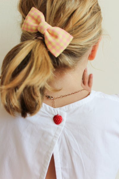 Woman with blonde hair tied in a ponytail with a pink checkered bow, wearing a white top with a red button.