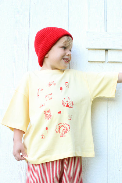 Child wearing yellow cotton t-shirt with hand-drawn orange childhood doodles on front and Long live childhood graphic on back