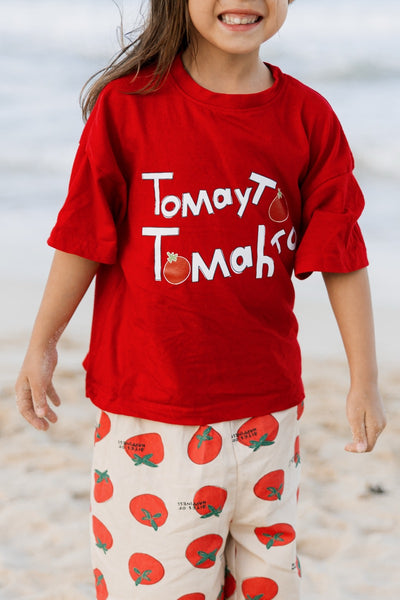Child wearing a red shirt with 'Tomato Tomatoes' text and matching pants on a beach.