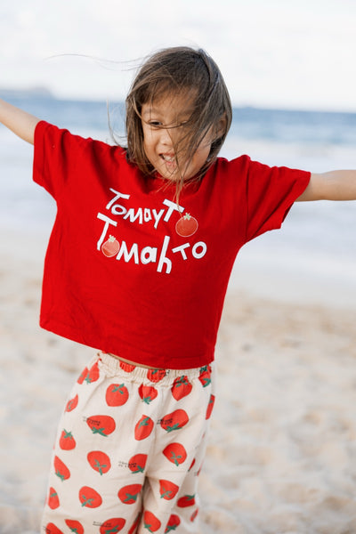 Child wearing a red tomato t-shirt with text and tomato-patterned pants on a beach.