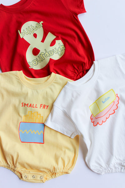 Red shirt with 'butter & sourdough' text, yellow romper with 'small fry' text, and white romper with 'butter dish' text on a light gray background.