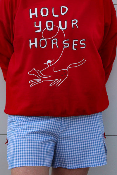 Woman wearing red sweatshirt with graphic that says hold your horses and wearing blue and white gingham shorts 