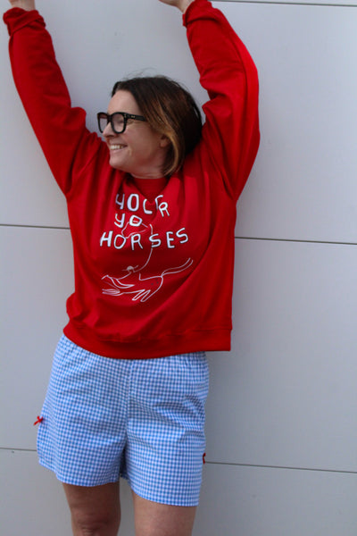 Woman wearing red sweatshirt with graphic that says hold your horses and wearing blue and white gingham shorts 